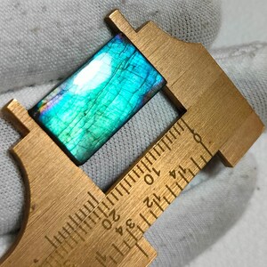 Könnte beinhalten: Ein rechteckiger Labradorit-Edelstein mit einer leuchtend blau-grün irisierenden Oberfläche, gemessen mit einem Messschieber aus Messing, der eine Länge von etwa 1,9 cm anzeigt. Der Stein hat eine rissige Textur und eine polierte Oberfläche.