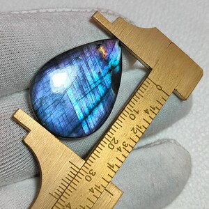 May include: A teardrop-shaped labradorite gemstone displaying iridescent blue and purple flashes, positioned against a brass caliper showing a measurement of approximately 3.2 centimetres in length along the stone's longest vertical axis.