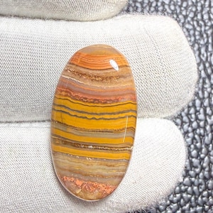 Könnte beinhalten: Ein ovaler Cabochon-Edelstein mit horizontalen Bändern in Senfgelb, gebranntem Orange, Hellbraun und Dunkelbraun. Der Stein hat eine glatte, polierte Oberfläche mit einem reflektierenden Glanz und einem ausgeprägten geschichteten Muster.