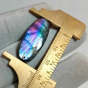 Puede incluir: Una piedra preciosa de labradorita de forma ovalada con destellos iridiscentes azules y morados, colocada junto a un calibrador de latón. La herramienta muestra que la piedra mide aproximadamente 3,2 cm de largo, con fisuras e inclusiones.