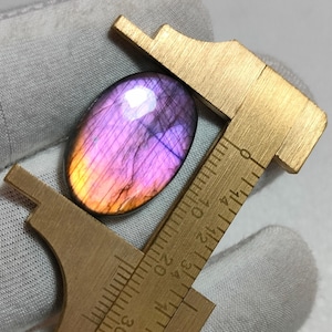May include: An oval-shaped labradorite gemstone with iridescent purple and orange hues, positioned within a brass caliper. The tool displays metric markings, showing the stone measures approximately 22 millimetres in length along its vertical axis.