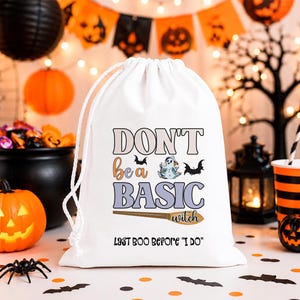 Könnte beinhalten: Ein weißer Beutel mit Kordelzug, der den Text DON'T be a BASIC witch und LAST BOO BEFORE I DO zeigt. Das Design umfasst ein kleines Gespenst, zwei schwarze Fledermäuse und eine braune Besen-Illustration auf der Vorderseite des Stoffbeutels.