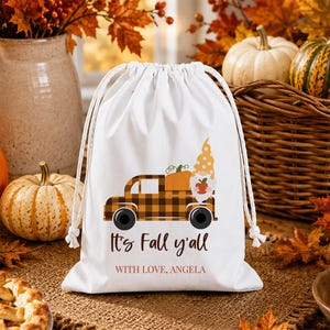 Könnte beinhalten: Ein weißer Beutel mit Kordelzug, der mit einem schwarzen und orangefarbenen karierten Lastwagen bedruckt ist, welcher einen Kürbis und einen Zwerg transportiert. Der Text lautet It's Fall y'all und WITH LOVE, ANGELA. Der Beutel hat weiße Kordeln.