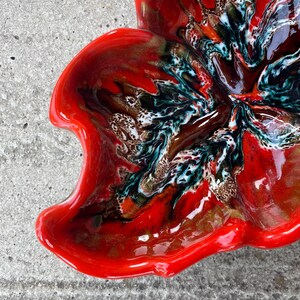 Op de afbeelding: Een glanzende keramische kom met een onregelmatige, organische vorm. Het oppervlak is voorzien van een levendig rood glazuur met wervelende, gemarmerde patronen in diepzwart, wit en groenblauw, wat een contrastrijk en vloeiend effect creëert.