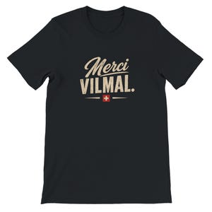 Könnte beinhalten: Ein schwarzes Kurzarm-T-Shirt mit dem beigen Schriftzug "Merci VILMAL." auf der Brust. Unter dem Text befindet sich ein kleines rotes Quadrat mit einem weißen Schweizer Kreuz, das von zwei horizontalen beigen Linien flankiert wird.