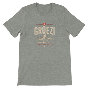 Könnte beinhalten: Ein graues, meliertes Kurzarm-T-Shirt mit einem Grafikdruck, der den Schriftzug GRÜEZI, ein kleines Schweizer Flaggensymbol, eine Bergillustration sowie den Text SWISS FOR HELLO mit einem kleinen Kreuzsymbol unter dem Bergmotiv zeigt.