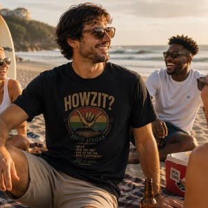 Könnte beinhalten: Ein schwarzes Kurzarm-T-Shirt mit einem Aufdruck, der den Text HOWZIT? über einer Shaka-Handgeste in einem sonnenuntergangsfarbenen Kreis zeigt. Unter dem Kreis steht der Text SOUTH AFRICAN for: HELLO, HOW ARE YOU, NICE TO SEE YOU, WHAT'S HAPPENING.