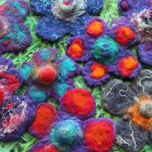 Op de afbeelding: Close-up van verschillende handgemaakte vilten bloemen in verschillende kleuren. De bloemen hebben een gelaagd ontwerp met levendige tinten paars, blauw, rood, oranje en blauwgroen. De vilten bloemen zijn gerangschikt op een groene ondergrond.