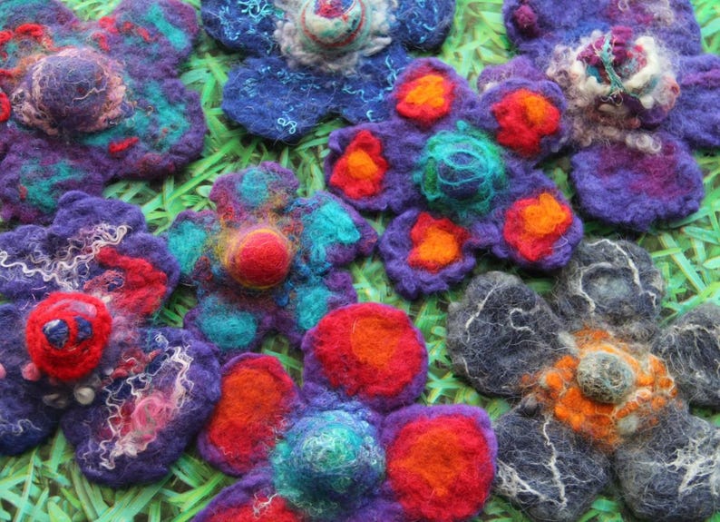 May include: Close-up of several handmade felt flowers in various colors. The flowers have a layered design with vibrant hues of purple, blue, red, orange, and teal. The felt flowers are arranged on a green surface.