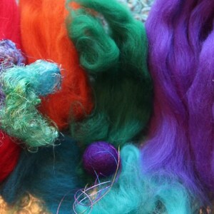 May include: A close-up of colorful fiber art materials. The image features vibrant hues of red, orange, green, teal, and purple. The materials appear to be wool or similar fibers, possibly for felting or other textile crafts.