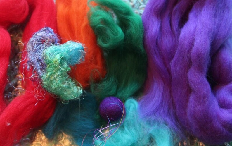May include: A close-up of colorful fiber art materials. The image features vibrant hues of red, orange, green, teal, and purple. The materials appear to be wool or similar fibers, possibly for felting or other textile crafts.