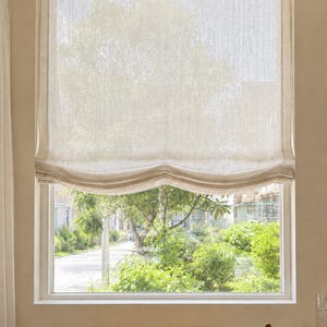 May include: A semi-sheer, off-white Roman shade made of a textured fabric hangs over a window. The shade features a soft, scalloped bottom edge and is partially raised, allowing natural light to filter through the weave into the interior space.