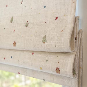 Puede incluir: Un estor romano beige de tela texturizada con un patrón repetitivo de pequeñas ilustraciones coloridas, que incluyen pinos verdes, casas marrones y varias estrellas rojas, azules y amarillas dispersas por la superficie.