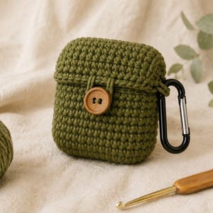 Könnte beinhalten: Eine olivgrüne, gehäkelte Schutzhülle für kabellose Ohrhörer mit strukturiertem Strickmuster, einem zentralen braunen Holzknopfverschluss und einem schwarzen Metallkarabiner an der Seite zur sicheren Befestigung und zum einfachen Transport.