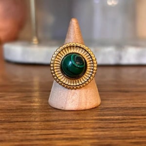 Op de afbeelding: Een goudkleurige ring met een ronde, donkergroene malachietsteen met natuurlijke banden. De steen is gezet in een brede, getextureerde goudkleurige zetting die een zonachtig patroon heeft met stralende lijnen rondom het midden.