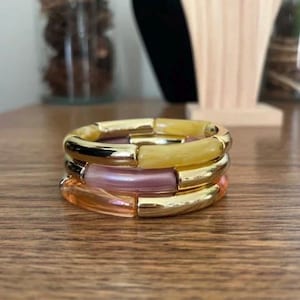 Peut inclure: Un empilement de trois bracelets circulaires composés de segments alternés en métal doré poli et en résine lisse translucide. Les segments en résine sont colorés dans des tons jaune pâle, rose tendre et une teinte orange translucide.