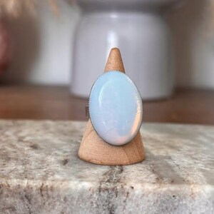 Peut inclure: Une bague ornée d'une grande pierre ovale blanc laiteux, lisse, présentant des reflets irisés bleus et roses. La pierre est montée sur un anneau métallique simple et présentée sur un support conique en bois. La surface est translucide.