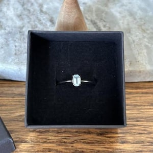 Peut inclure: Une bague argentée ornée d'une pierre précieuse unique, transparente et taillée en émeraude, montée sur un anneau métallique fin. La bague est présentée au centre d'un écrin carré doublé de velours noir, offrant un contraste saisissant.