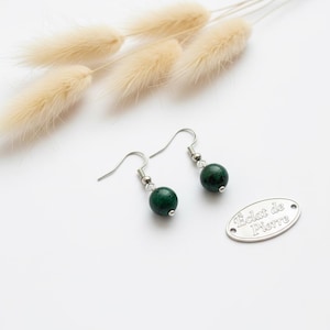 May include: A pair of drop earrings featuring round, dark green mottled stone beads suspended from silver-toned metal hooks. Beside the earrings lies a small, oval-shaped silver-toned metal tag engraved with the text Éclat de Pierre.