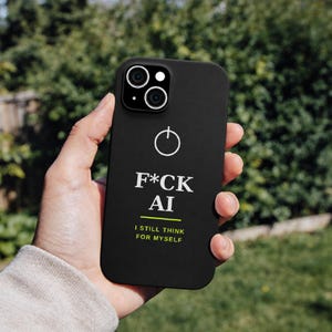 Peut inclure: Une coque de smartphone noir mat arborant une icône de bouton d'alimentation blanche en haut. Sous l'icône, le texte F*CK AI est imprimé en blanc, suivi d'une ligne horizontale vert citron et du texte I STILL THINK FOR MYSELF en vert citron.