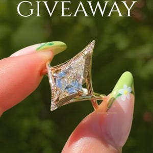 May include: A gold ring featuring a large kite-shaped center stone with a faceted surface, surrounded by a delicate pavé-set border. The band has a unique geometric design with sharp, angular points extending from the central setting.