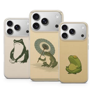May include: Three smartphone cases featuring frog illustrations. The left case shows a dark green frog with a bowl. The center case displays a green frog holding a blue-green parasol. The right case features a light green frog sitting in a meditative pose.