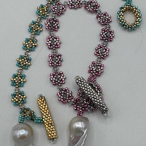 May include: A beaded necklace with two large white pearls, one on each end. The necklace is made of small, colorful beads in shades of teal, gold, pink, and silver. The beads are arranged in a pattern of small squares.
