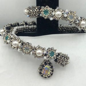 Peut inclure: Un bracelet en perles argentées et noires avec des perles blanches et des pierres précieuses vertes. Le bracelet a un charme suspendu avec une pierre précieuse irisée.
