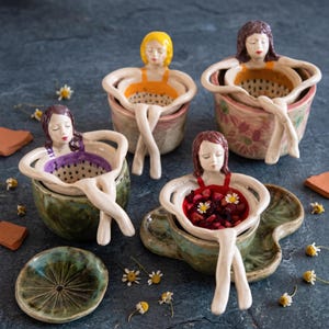 May include: Four ceramic tea strainers shaped like figures sitting in bowls. Each figure has a different hair colour and swimsuit shade, including yellow, brown, purple, and red. The strainers feature perforated bases and are resting on matching ceramic cups.