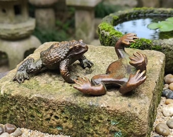 May include: Two bronze-coloured frog figurines resting on a textured, mossy stone block. One frog is positioned in a crouched, upright stance, while the other is lying on its back with its limbs extended upwards, showcasing a detailed, bumpy skin texture.