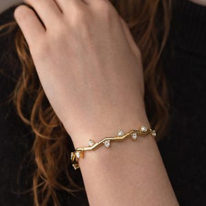 Puede incluir: Una pulsera de tono dorado con un diseño de eslabones metálicos en forma de zigzag o chevron. La pulsera está decorada con pequeñas piedras claras de corte redondo engastadas en cada ángulo exterior de la estructura geométrica.