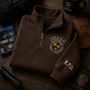 May include: A brown quarter-zip pullover featuring an embroidered S.T.A.R.S. Dept Raccoon Police circular patch on the chest with three yellow stars. The left sleeve cuff displays the white embroidered text R.P.D. The garment has a metal zipper.