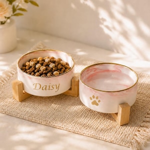 May include: Two round ceramic pet bowls with a pink marble pattern and gold rims, each resting on a light wooden stand. One bowl is filled with brown kibble and features the name Daisy in gold script, while the other contains water and a gold paw print.