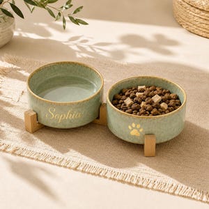 May include: Two sage green ceramic pet bowls set on a wooden stand. One bowl is filled with water and features the name Sophia in gold script. The other bowl is filled with brown dry pet food and displays a gold paw print graphic on the side.