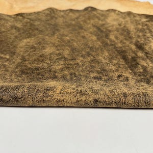 Op de afbeelding: Een stuk leer met een donkerbruine en zwarte gevlekte, verweerde oppervlaktestructuur. Het materiaal heeft een omgevouwen rand die een lichtere, bruine en gladde onderkant onthult, wat het contrast tussen de twee zijden laat zien.