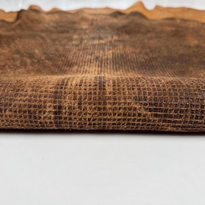 Op de afbeelding: Een stuk donkerbruin leer met een opvallende rasterachtige reliëftextuur. Het materiaal is omgevouwen om het textuuroppervlak te onthullen, met een diepbruine kleur en donkerdere schaduwen in de verdiepte delen van het rasterpatroon.