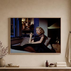 May include: A framed horizontal wall art print featuring a scene of a blonde person in a black dress holding a cigarette, sitting on a bed in a dimly lit room with blue window light, displayed in a neutral-toned room with a vase and towels.