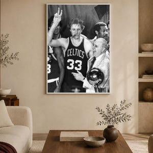 May include: A black and white framed photograph depicting a basketball player wearing a Celtics jersey with the number 33, holding up two fingers, with a cigar in their mouth, standing next to a person holding a trophy in a celebratory setting.