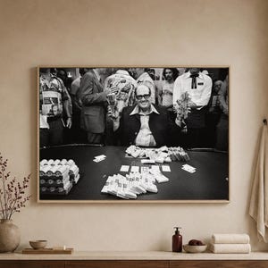 May include: A black and white framed photograph depicting a person wearing glasses and a dark jacket sitting at a casino table. The person holds stacks of cash in both hands, with additional piles of currency and playing cards spread across the table.