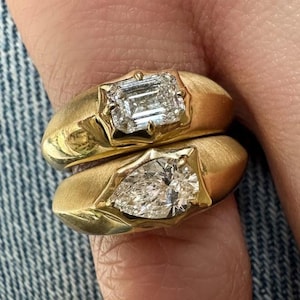 Peut inclure: Deux bagues en or à la finition mate, chacune ornée d'une pierre précieuse à facettes. La bague supérieure porte une pierre rectangulaire taille émeraude, tandis que la bague inférieure présente une pierre en forme de poire, serties sur griffes.