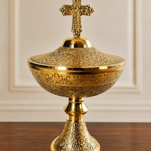 May include: A gold-toned metal ciborium featuring an ornate cross finial on the lid. The bowl and base are decorated with intricate floral engravings and leaf patterns, with a polished finish on the smooth sections and a textured, dark inlay.