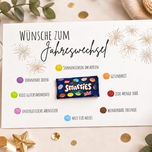 Könnte beinhalten: Eine weiße rechteckige Karte mit dem Text Wünsche zum Jahreswechsel und goldenen Feuerwerksillustrationen. Ein kleines blaues Smarties-Tütchen ist in der Mitte befestigt, umgeben von bunten Punkten und deutschen Phrasen wie Sonnenschein im Herzen.