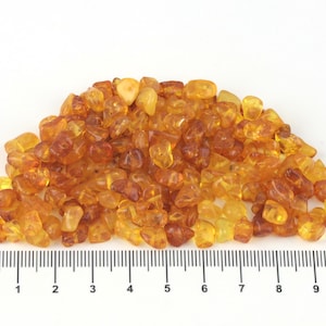 Può includere: Un mucchio di pietre d'ambra lucidata in varie tonalità di giallo e marrone. Le pietre hanno forme e dimensioni irregolari, con un aspetto traslucido. Un righello è posto sotto per la scala.