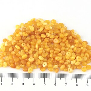 May include: A collection of polished amber beads in shades of yellow and orange. The small beads, with varied shapes, are pre-drilled for stringing. A ruler at the base provides measurements in centimetres.