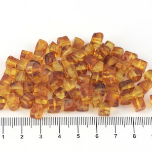 May include: A pile of amber-colored, cube-shaped beads. The beads have a translucent quality, with variations in color from light yellow to deep brown. A ruler is placed below the beads, showing measurements in inches.