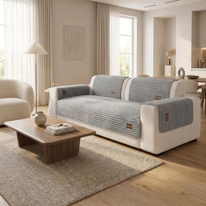 May include: A light gray, ribbed sofa cover draped over a cream-colored couch. The cover features a textured, horizontal striped pattern and includes small, rectangular brown fabric labels with the text "hometex" attached to the armrests and back.
