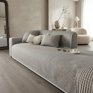May include: A light gray sofa featuring a textured waffle-knit fabric cover. The sofa includes a rectangular backrest, a long seat cushion, and several decorative throw pillows in white and gray tones, arranged neatly along the back of the furniture.