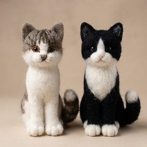 Könnte beinhalten: Zwei nadelgefilzte Katzenfiguren sitzen nebeneinander. Die linke Katze ist grau-weiß getigert mit braunen Augen. Die rechte Katze ist schwarz-weiß mit weißer Brust und weißem Gesichtsfleck. Beide haben spitze Ohren, kleine Nasen und geringelte Schwänze.