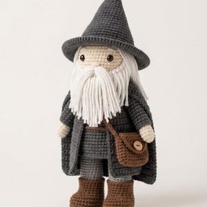 Könnte beinhalten: Eine gehäkelte Zaubererfigur mit einem dunkelgrauen spitzen Hut, einem passenden langen Umhang und einer Tunika. Die Figur hat einen langen weißen Garnbart, helle Hände, braune Stiefel und eine kleine braune Umhängetasche mit einem Holzknopf.