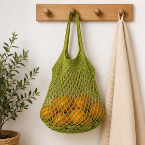 May include: A bright green crochet market bag with a diamond-shaped mesh pattern, featuring two sturdy handles and filled with several round, orange citrus fruits. The bag is suspended from a wooden wall hook, showcasing its flexible, open-weave design.